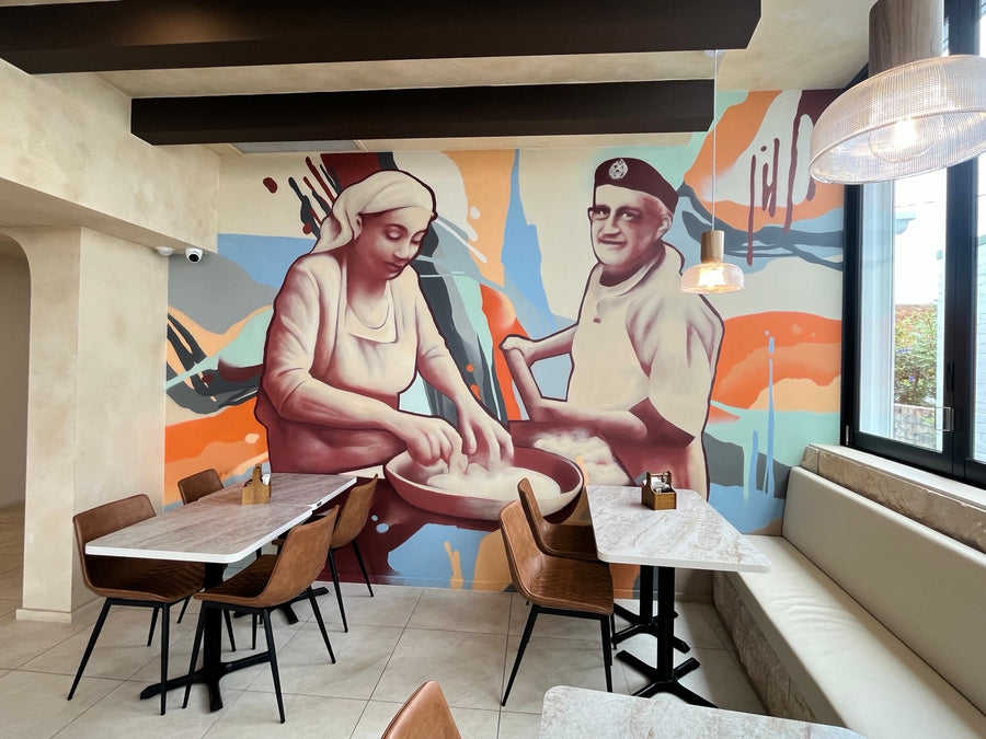 Colourful DMuly café mural of a woman and man preparing dough, displayed behind restaurant tables.